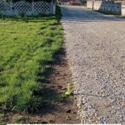 Wytyczanie granic działki: żółty palik wbity w ziemię, oznaczający granicę między trawnikiem a żwirową drogą, z widokiem na domy i betonowe ogrodzenie.