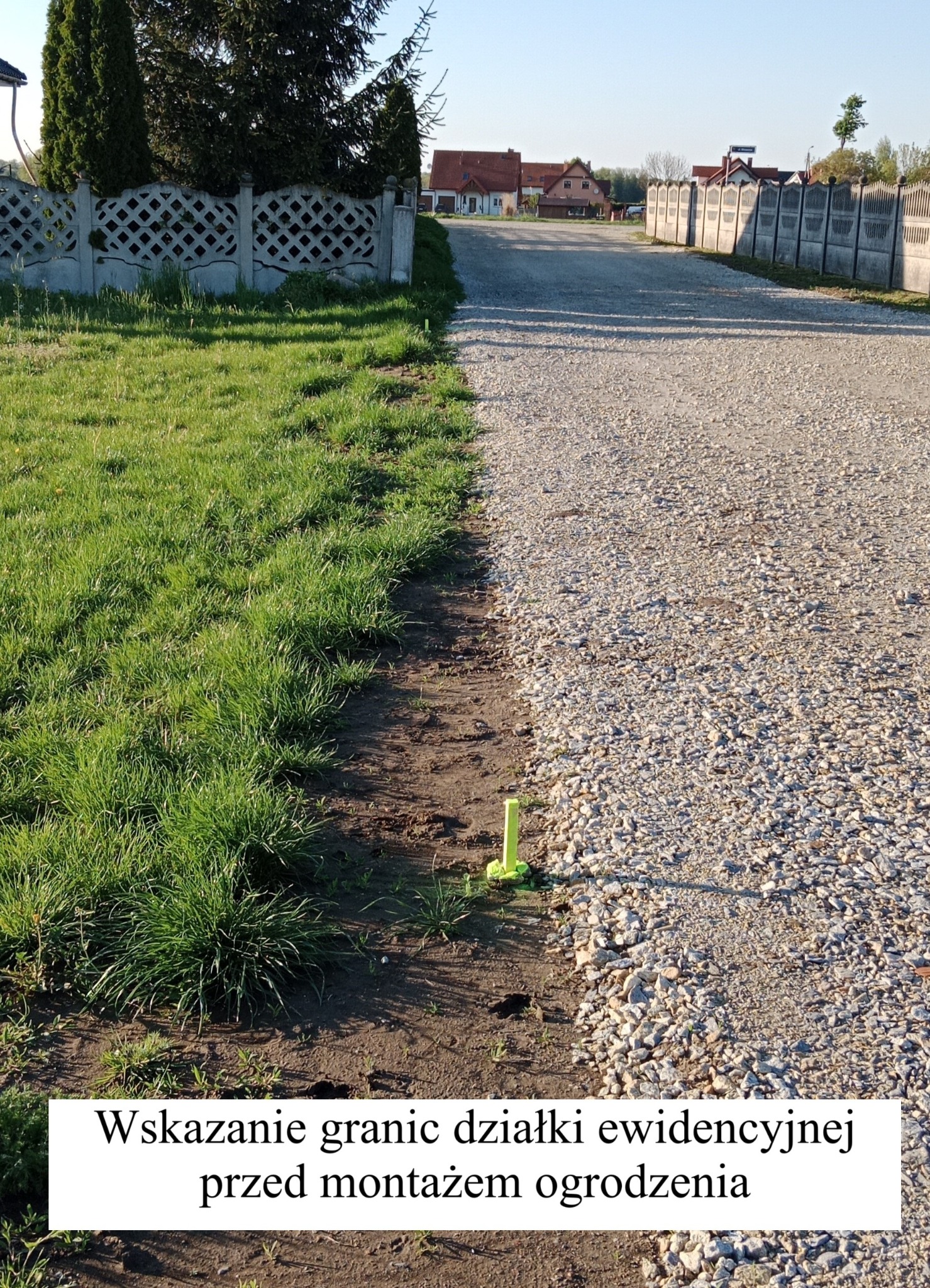 Wytyczanie granic działki: żółty palik wbity w ziemię, oznaczający granicę między trawnikiem a żwirową drogą, z widokiem na domy i betonowe ogrodzenie.