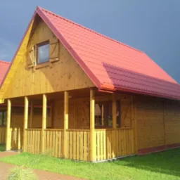 Drewniany domek letniskowy z czerwonym dachem na tle ciemnego nieba. Widoczna weranda z barierkami i numer domu. Ujęcie z poziomu trawnika.