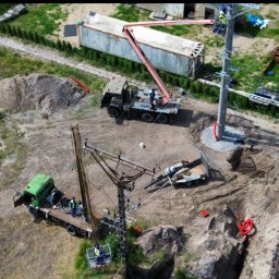 ALTA-Instal Tomasz Chodnicki - Montaż słupa energetycznego z użyciem dźwigu i platformy roboczej. Widok z góry na plac budowy z maszynami i pracownikami w kaskach.