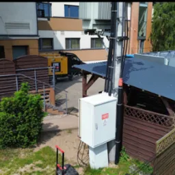 Biała skrzynka elektryczna z ostrzeżeniem na słupie, kable i budynki w tle. Widoczny generator prądu. Ujęcie z góry, w dzień.