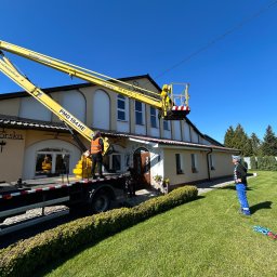 ALTA-Instal Tomasz Chodnicki - Pracownik na podnośniku koszowym, prawdopodobnie podczas montażu instalacji odgromowej na budynku. Widoczne przewody elektryczne i budynek z ogrodem.