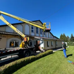 Pracownik na podnośniku koszowym, prawdopodobnie podczas montażu instalacji odgromowej na budynku. Widoczne przewody elektryczne i budynek z ogrodem.
