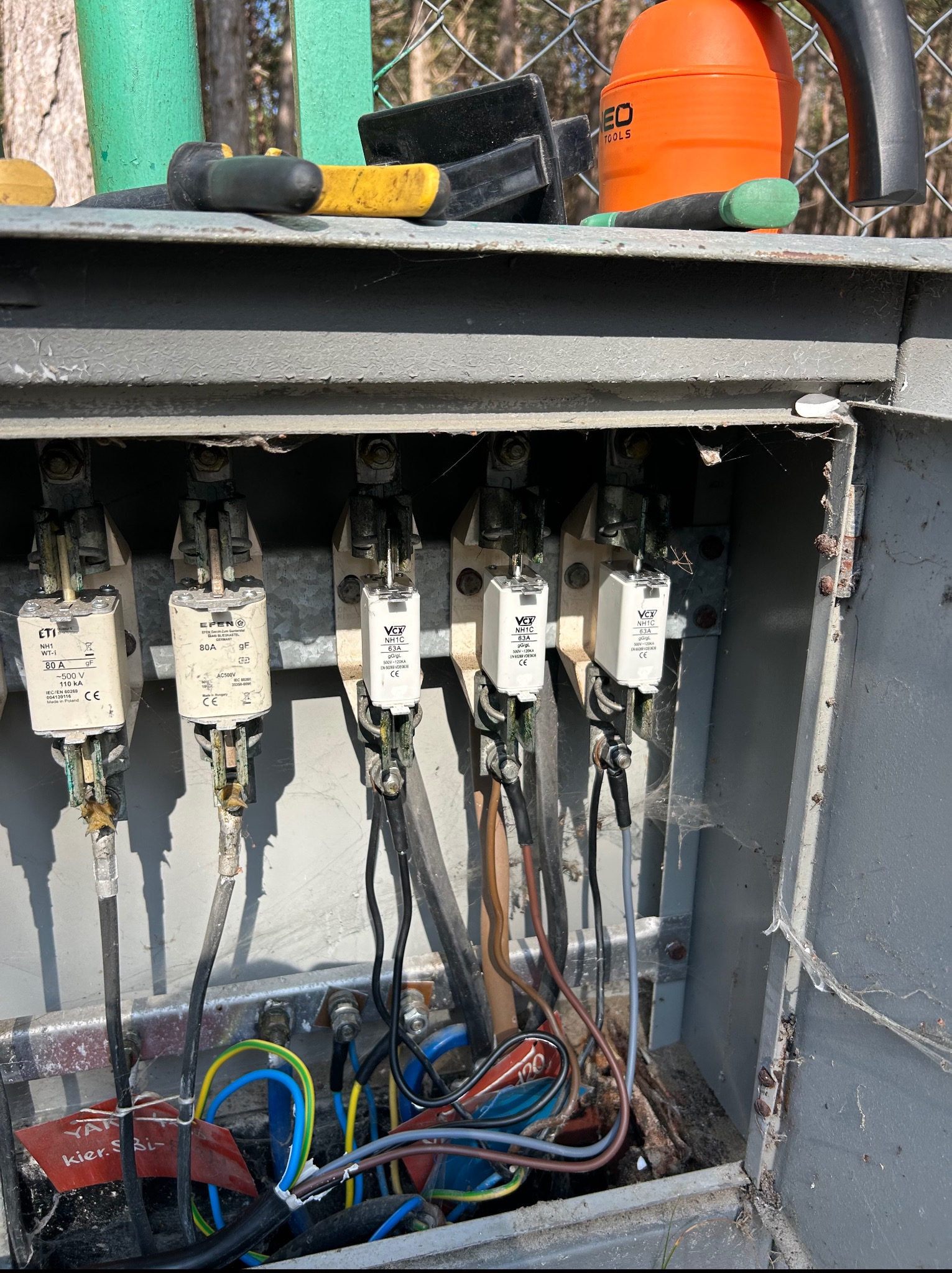 Otwarta skrzynka elektryczna z bezpiecznikami i przewodami. Widoczne narzędzia na górze skrzynki. Detale: zabrudzenia i pajęczyny wewnątrz skrzynki, etykieta na przewodach.