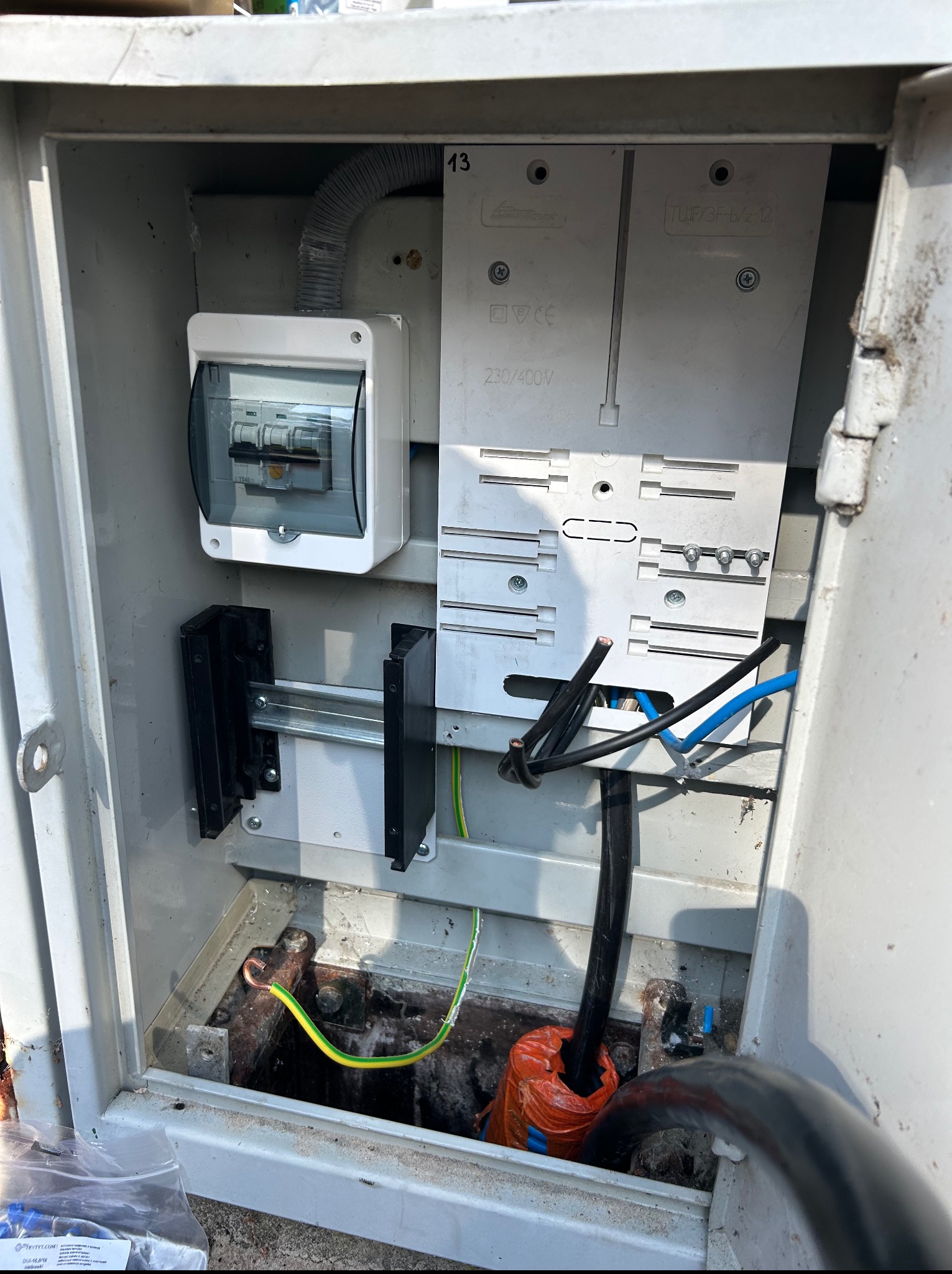 Otwarta skrzynka elektryczna z zamontowanym bezpiecznikiem, listwami DIN i przewodami. Widoczne złącza kablowe i oznaczenia napięcia 230/400V. Elementy instalacji elektrycznej.