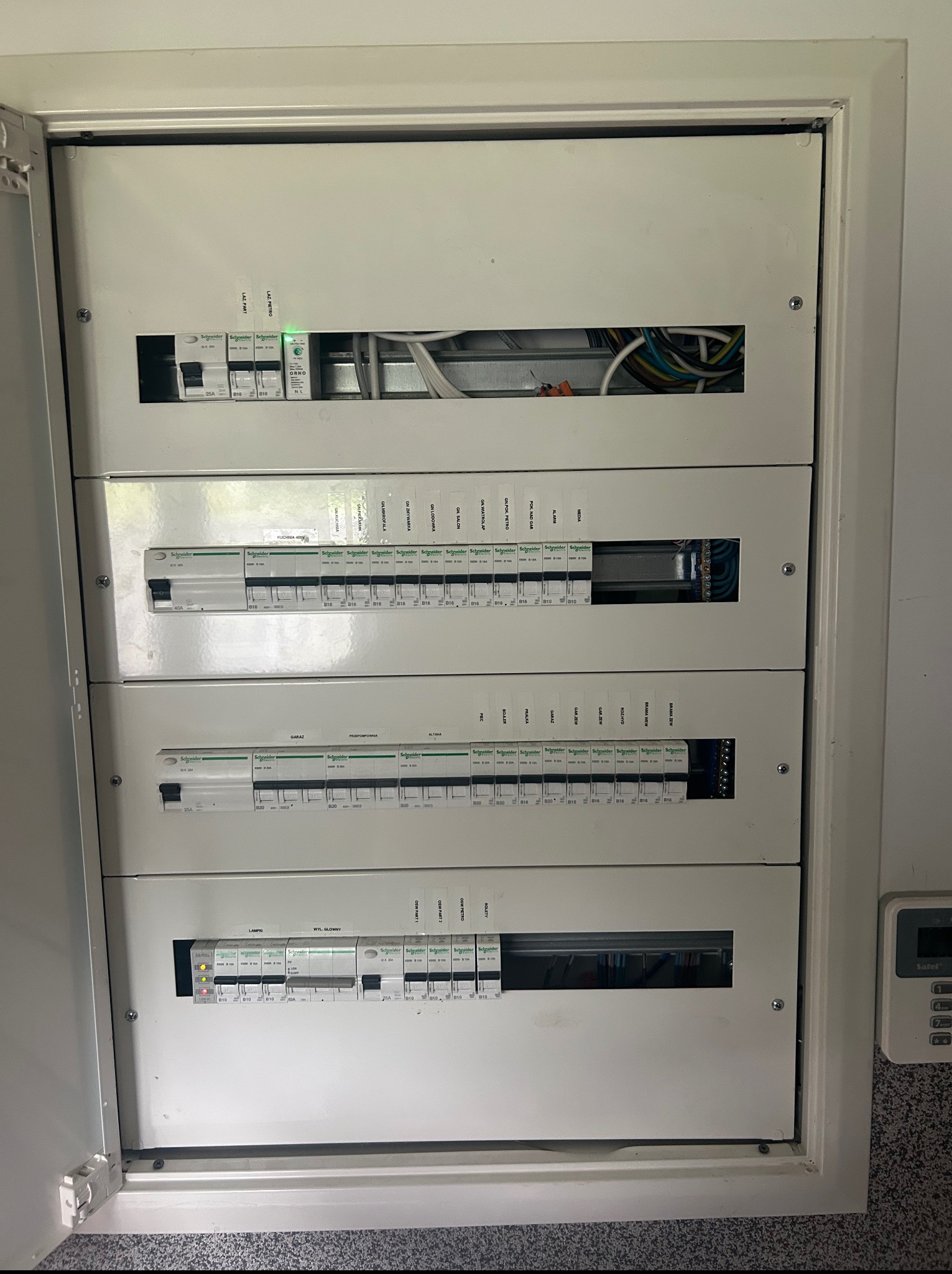 Otwarta skrzynka elektryczna z bezpiecznikami Schneider Electric, widoczne przewody i oznaczenia obwodów, w tle panel sterowania alarmem.