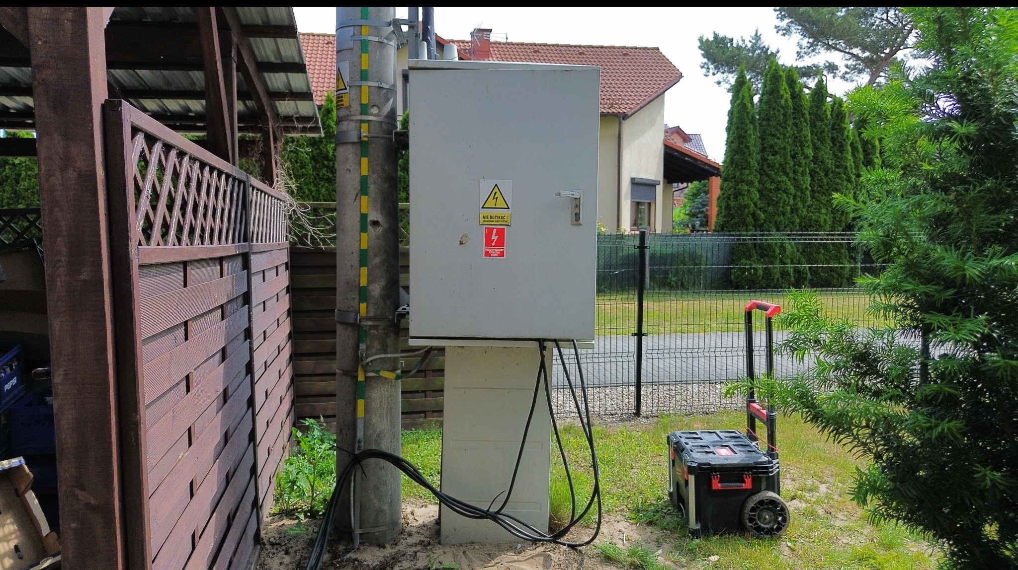 Szara skrzynka elektryczna z ostrzeżeniami na słupie, obok narzędzia w skrzynce na kółkach. Widoczne kable, trawa i ogrodzenie. Dom w tle.