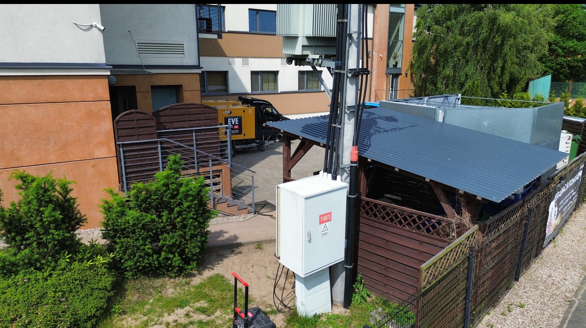 Biała skrzynka elektryczna z ostrzeżeniem na słupie, kable i budynki w tle. Widoczny generator prądu. Ujęcie z góry, w dzień.