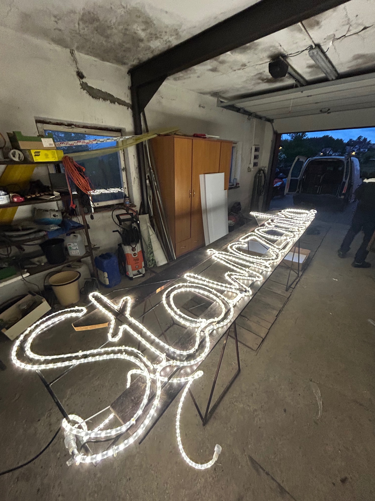 Konstrukcja świetlna z napisem ułożona na stole w warsztacie, widoczny fragment sylwetki osoby w tle i otwarte drzwi garażowe.