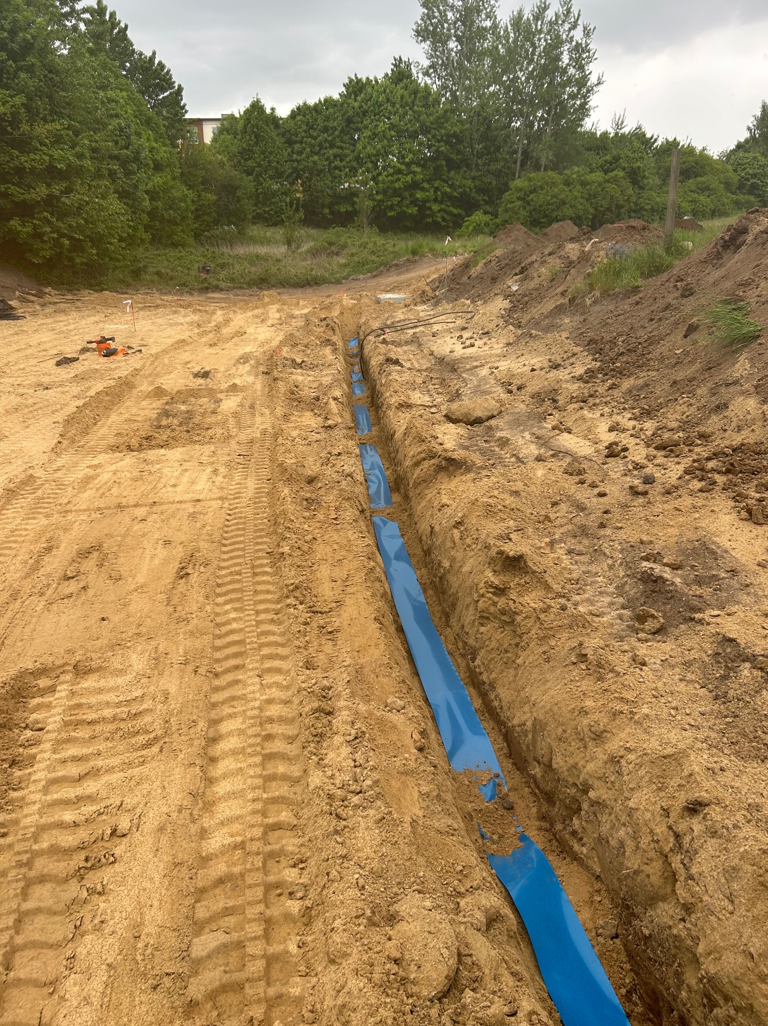 Wykop z niebieską rurą w ziemi, prawdopodobnie pod instalację elektryczną. Widoczne ślady opon i narzędzia w tle. Prace ziemne w trakcie realizacji, teren przygotowany.