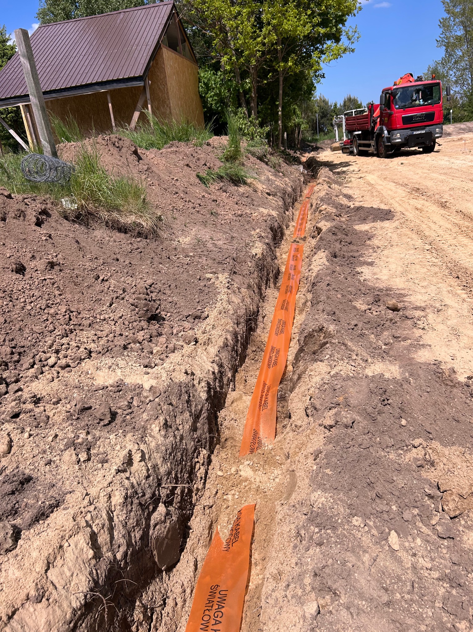Wykopany rów z pomarańczową osłoną kabla światłowodowego. W tle budynek w trakcie budowy i czerwona ciężarówka. Widok z góry.
