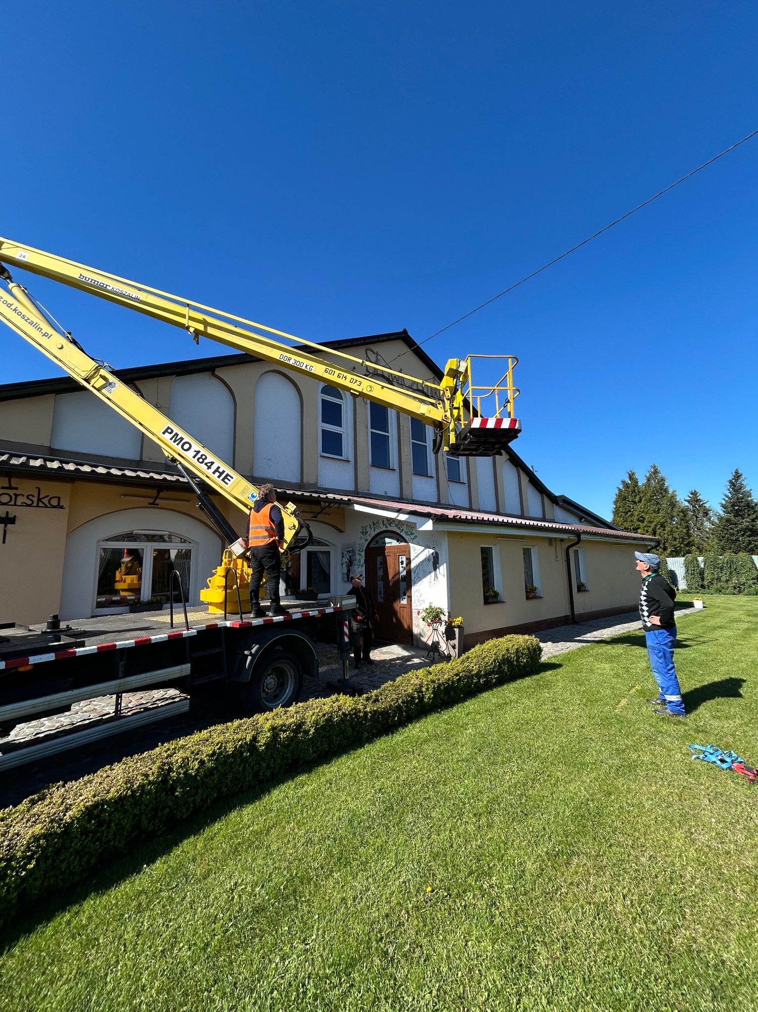 Pracownik na podnośniku koszowym, prawdopodobnie podczas montażu instalacji odgromowej na budynku. Widoczne przewody elektryczne i budynek z ogrodem.