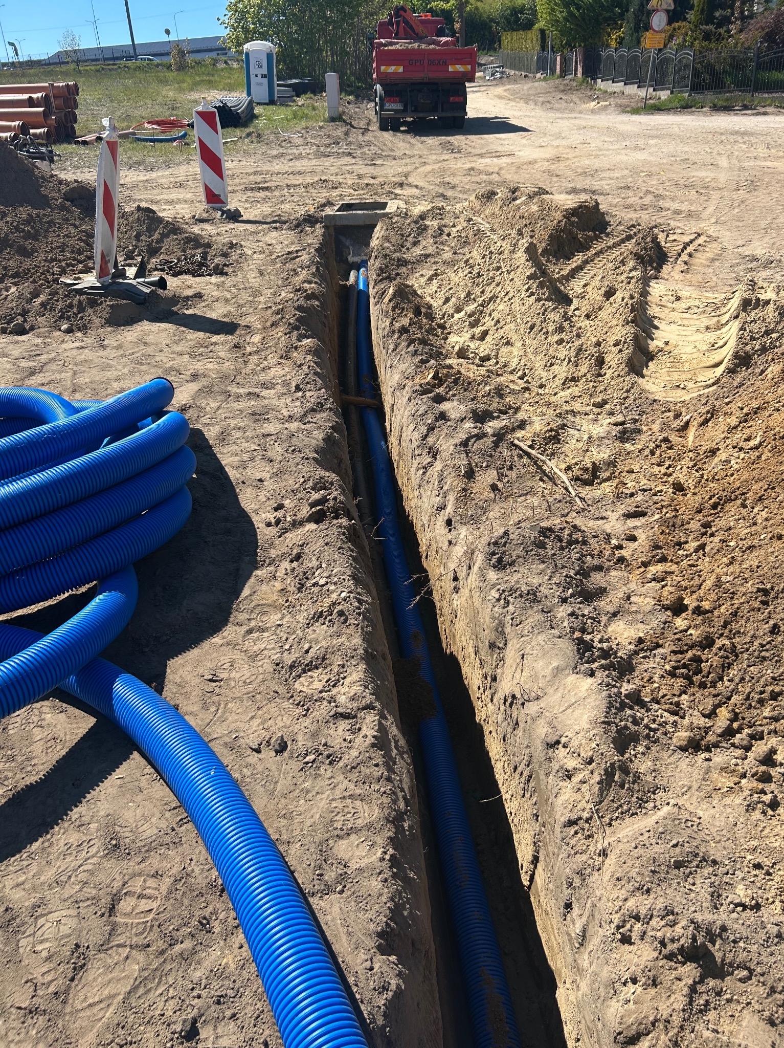Błękitne rury karbowane ułożone w wykopie ziemnym, widoczne z góry, z fragmentem ciężarówki w tle i elementami infrastruktury drogowej.