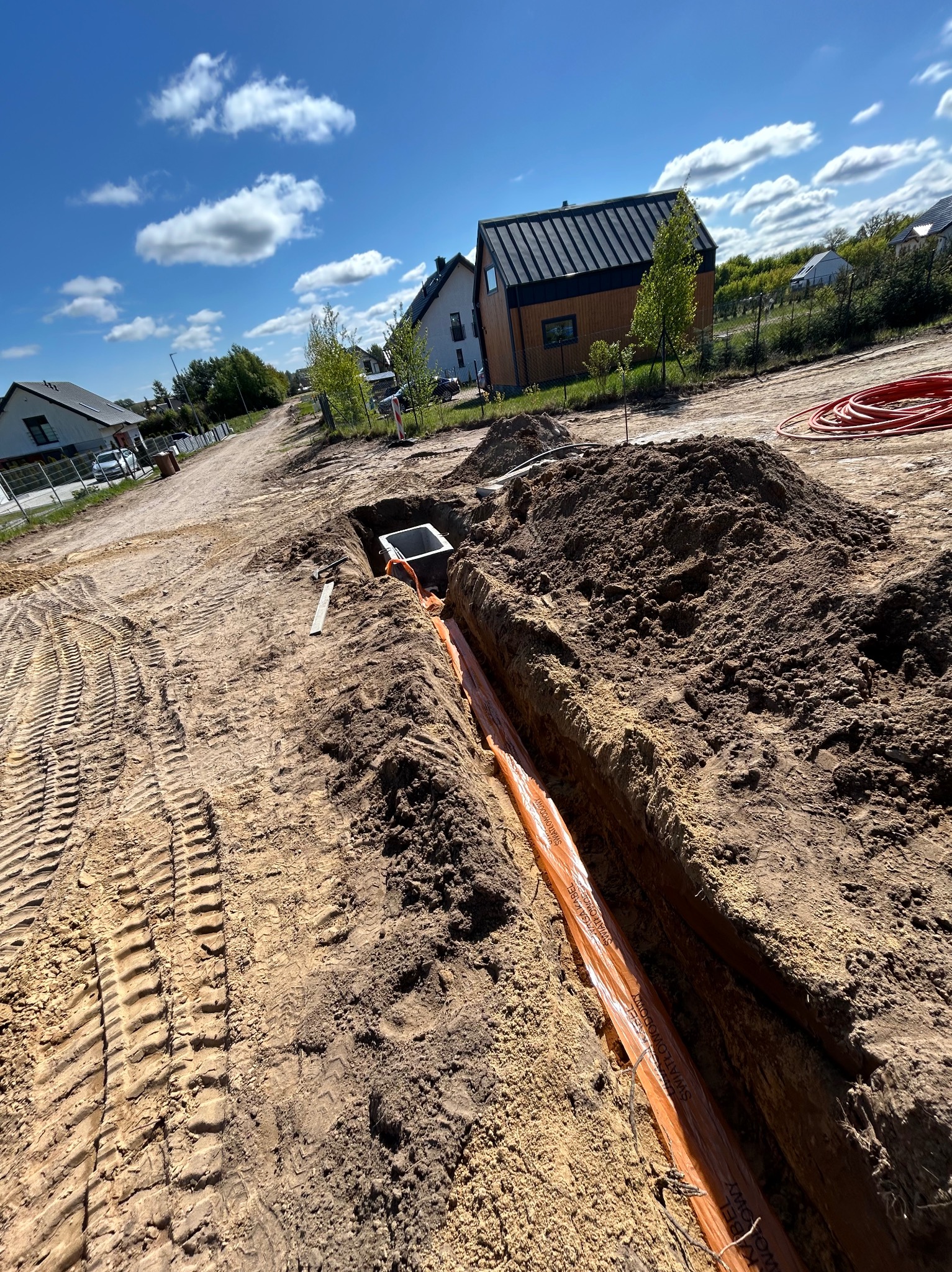 Wykop z pomarańczowymi kablami światłowodowymi w ziemi, widoczna studzienka kanalizacyjna i zabudowa w tle. Ślady opon na ziemi.