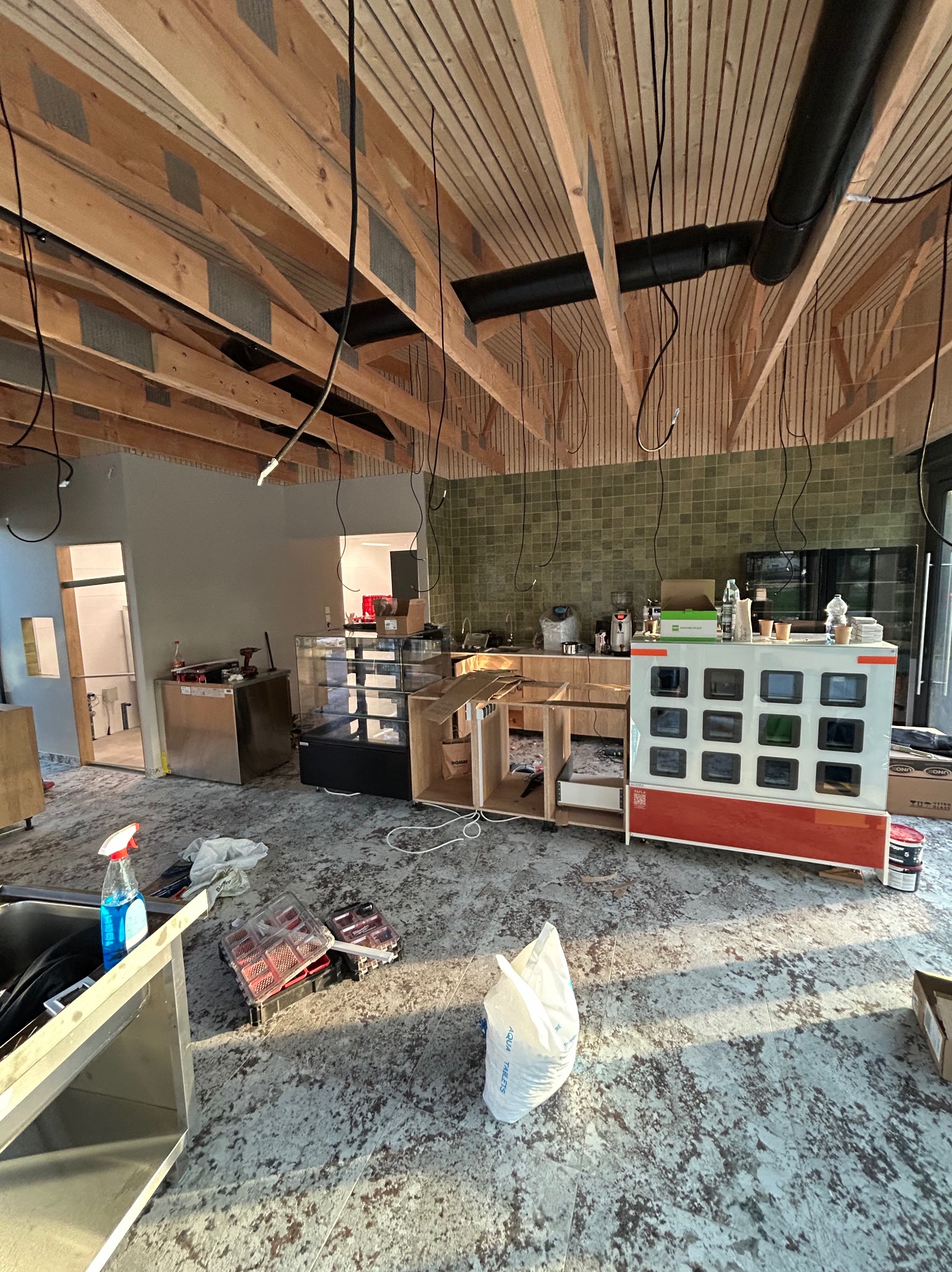 Wnętrze lokalu gastronomicznego w trakcie prac wykończeniowych: widoczne kable zwisające z sufitu, elementy wyposażenia baru i podłoga w trakcie układania.