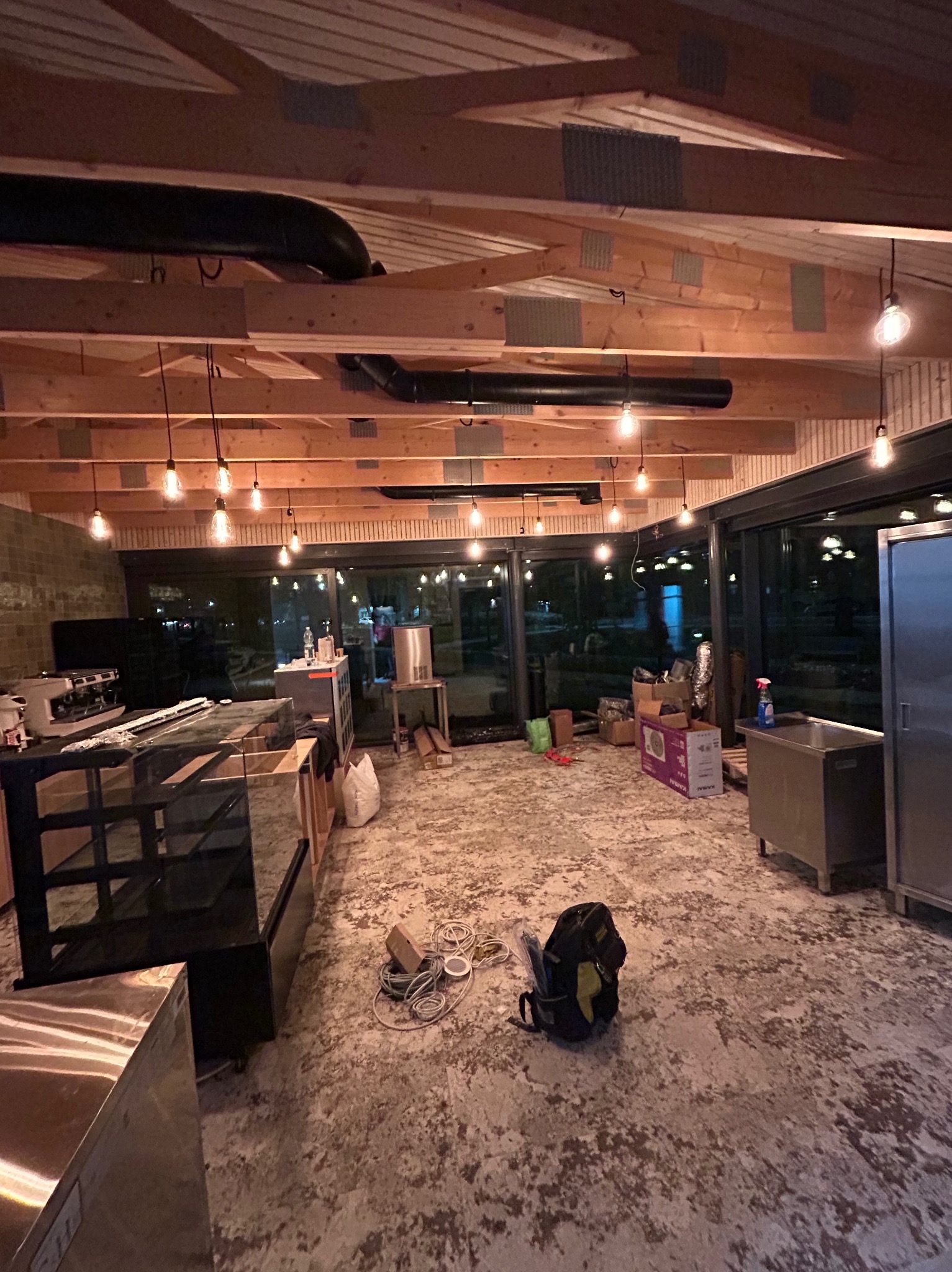 Wnętrze lokalu w trakcie prac wykończeniowych: widoczne elementy instalacji pod sufitem, surowa podłoga, sprzęt gastronomiczny i torba narzędziowa na posadzce.