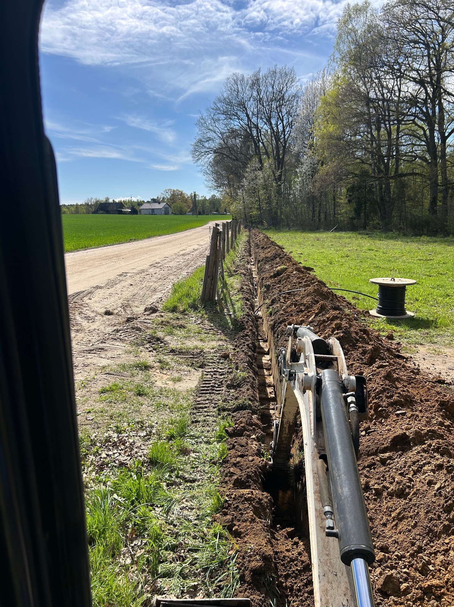 Koparka wykopuje wąski rów w ziemi wzdłuż ogrodzenia, rolka kabla w tle. Widok z kabiny operatora, słoneczny dzień.