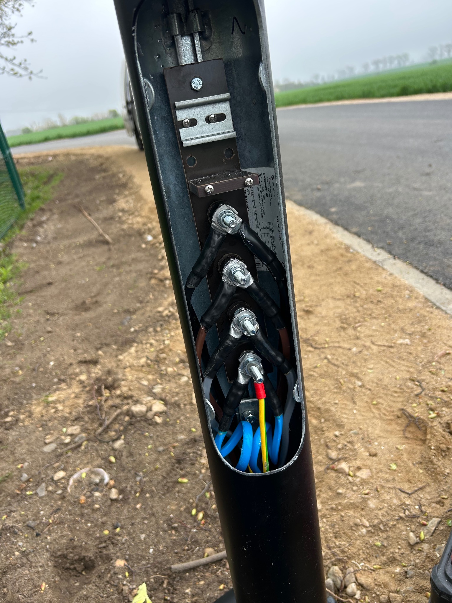 Otwarta obudowa słupa oświetleniowego z widocznymi, podłączonymi przewodami elektrycznymi w kolorach niebieskim, żółto-zielonym i czerwonym. Widoczne złącza i metalowe elementy mocujące.