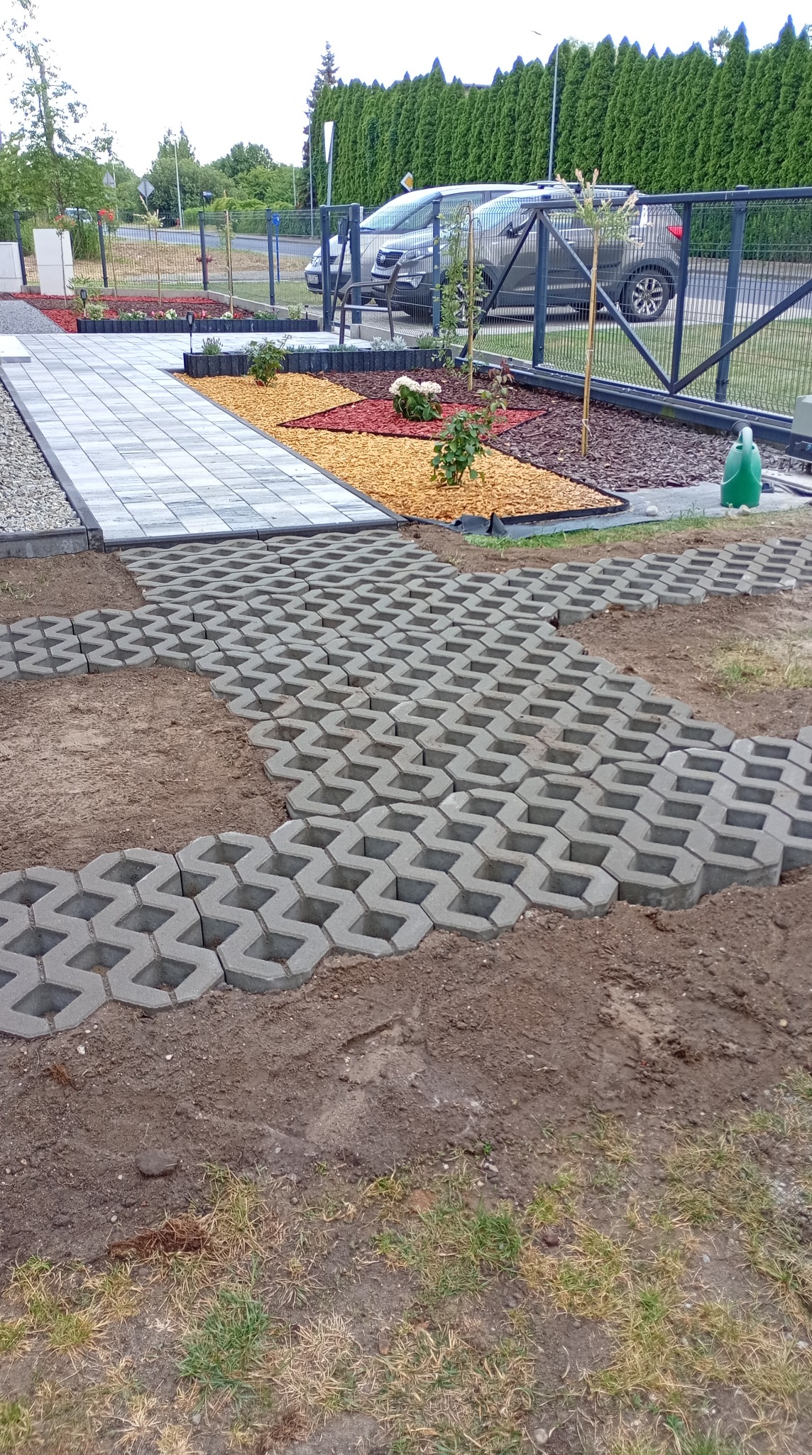 Układanie ażurowych płyt betonowych na ziemi, widoczne fragmenty ułożonego chodnika z kostki brukowej, w tle rabaty z korą i roślinami oraz ogrodzenie.