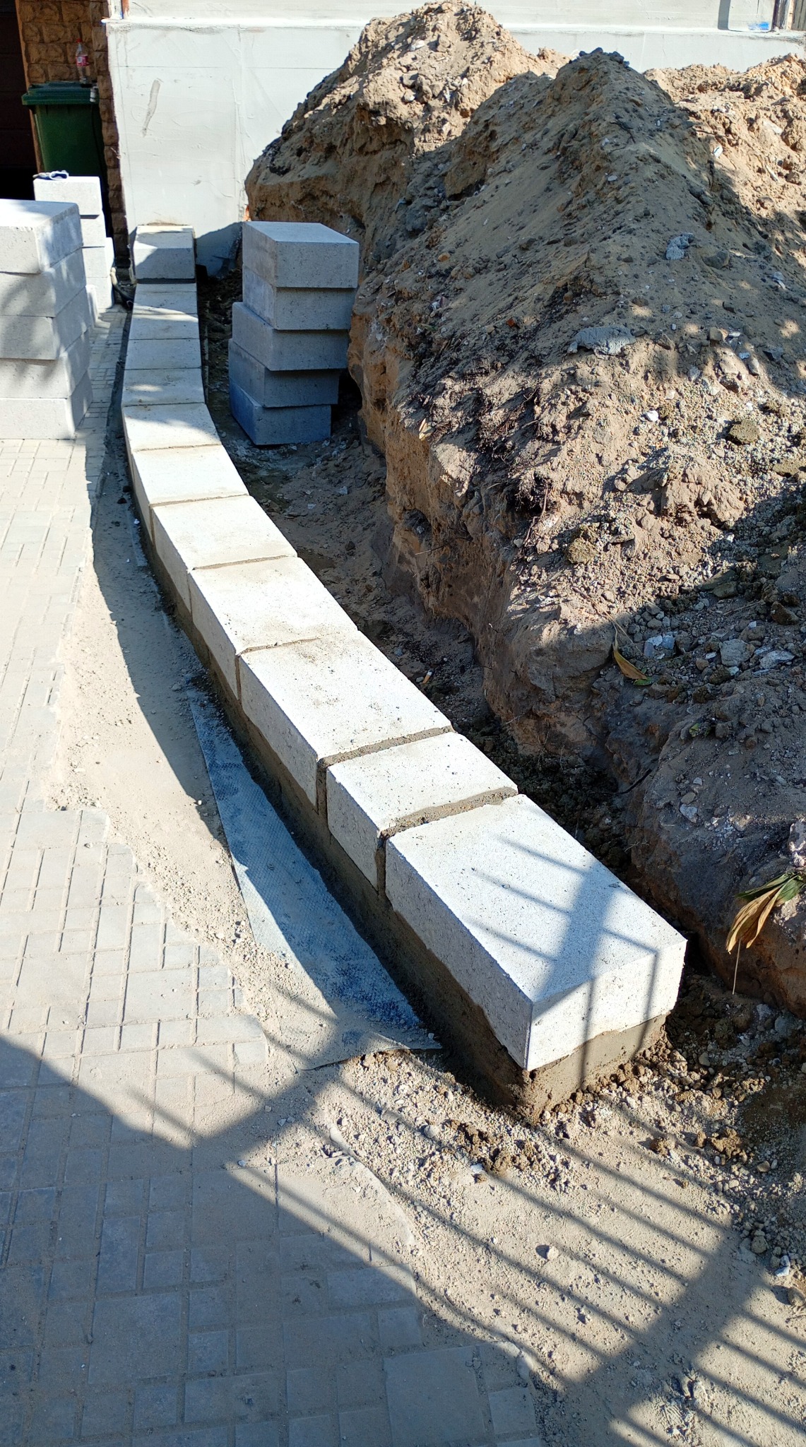 Układanie obrzeży z betonowych bloczków wzdłuż wykopu. Częściowo ułożone obrzeże z widoczną zaprawą. Stos bloczków obok, ziemia z wykopu w tle.