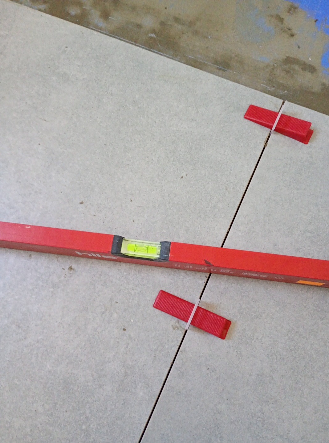 Ułożone jasne płytki podłogowe z czerwoną poziomicą i czerwonymi krzyżykami dystansowymi. Widok z góry na fugi i narzędzia glazurnicze.