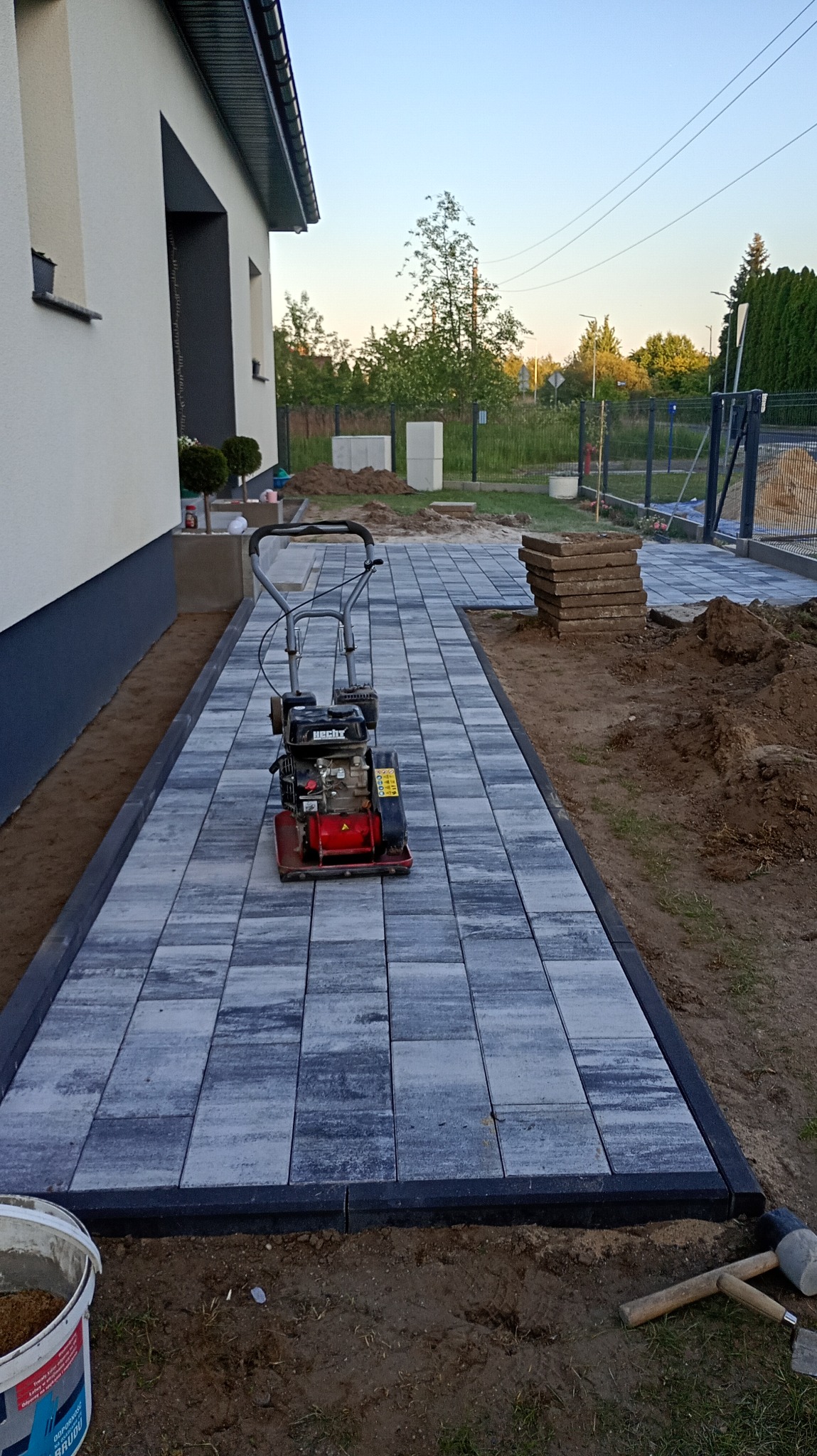 Układanie kostki brukowej na posesji domu jednorodzinnego. Widoczna ubijarka, ułożona część ścieżki z kostki w odcieniach szarości, obrzeża oraz narzędzia.