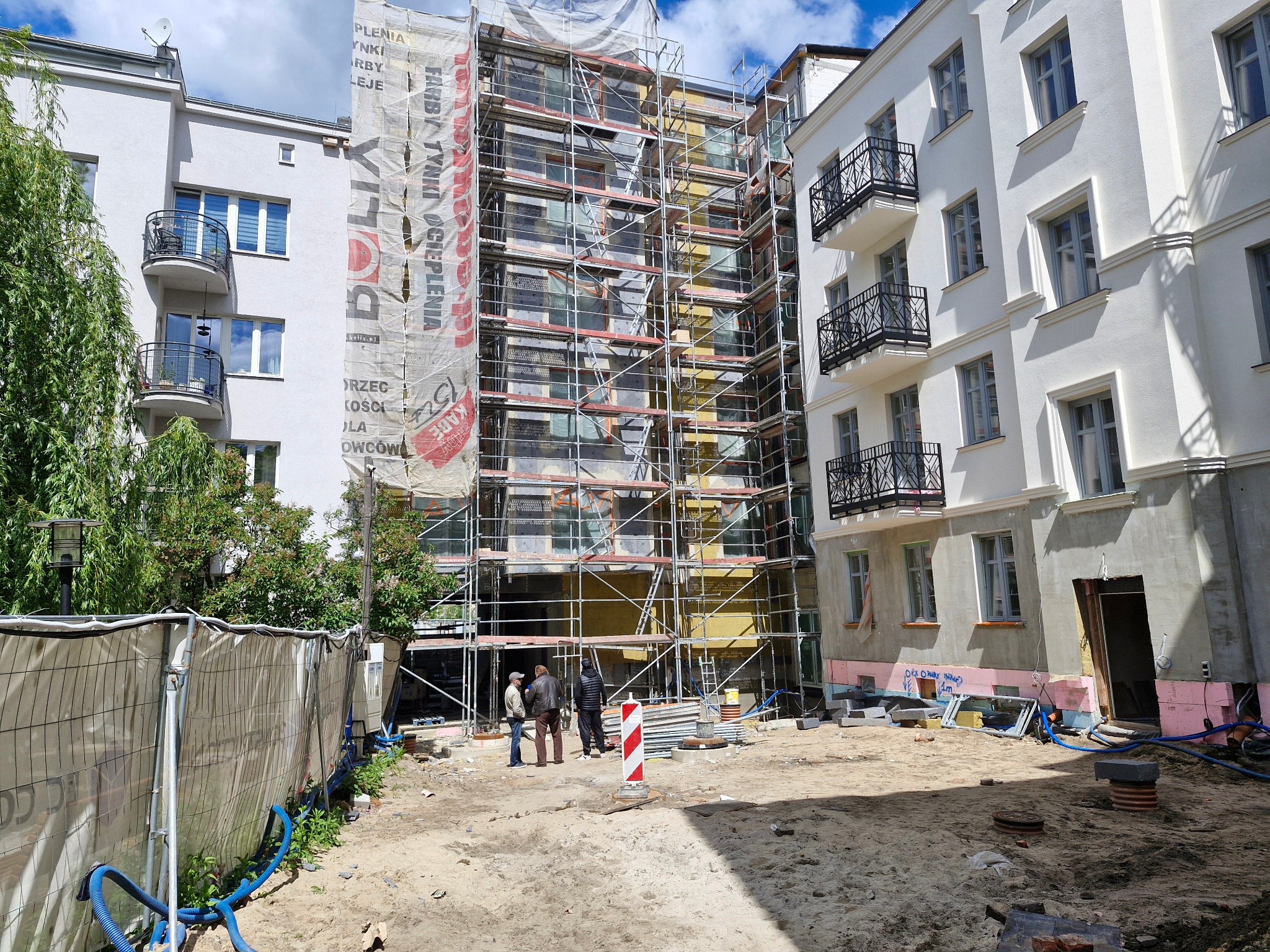 Prace elewacyjne w toku: rusztowania zasłonięte banerem reklamowym, ocieplenie budynku w trakcie. Widoczni pracownicy na placu budowy, przygotowany teren.