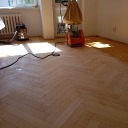 Strefa Parkietu - Proces renowacji parkietu dębowego w jodełkę. Widoczna cykliniarka Scorpion i odkurzacz. Część podłogi już odnowiona, jaśniejsza, reszta ciemniejsza.