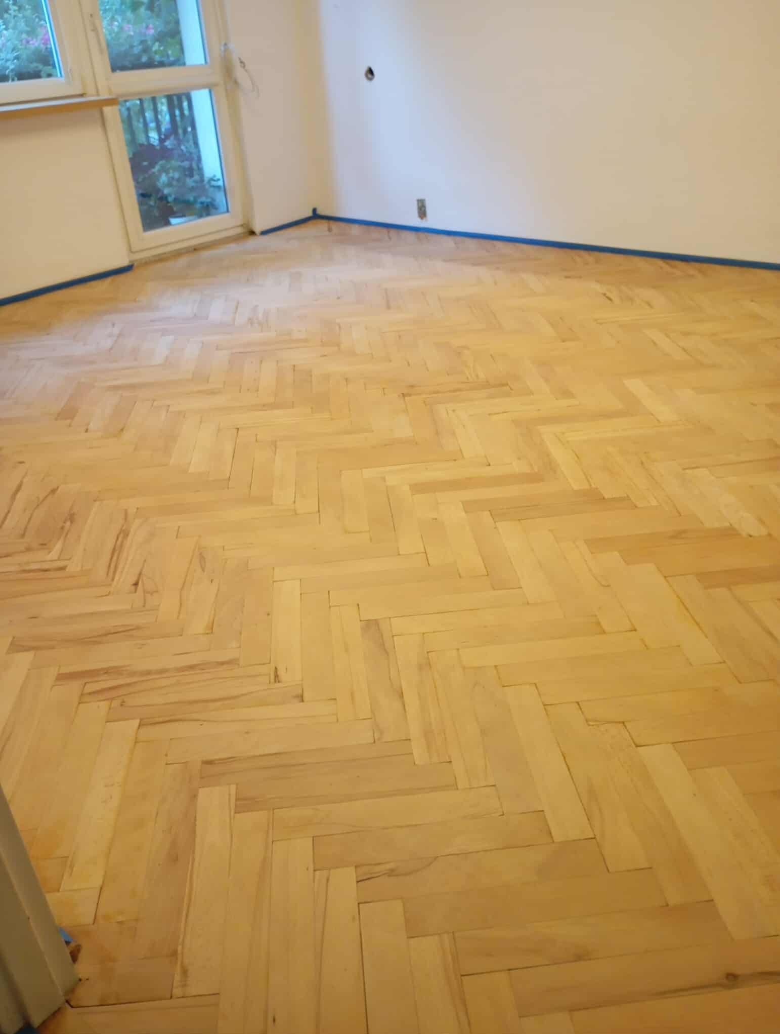 Jasny parkiet bukowy ułożony w jodełkę po cyklinowaniu, zabezpieczony niebieską taśmą malarską przy ścianach. Widok na okno.