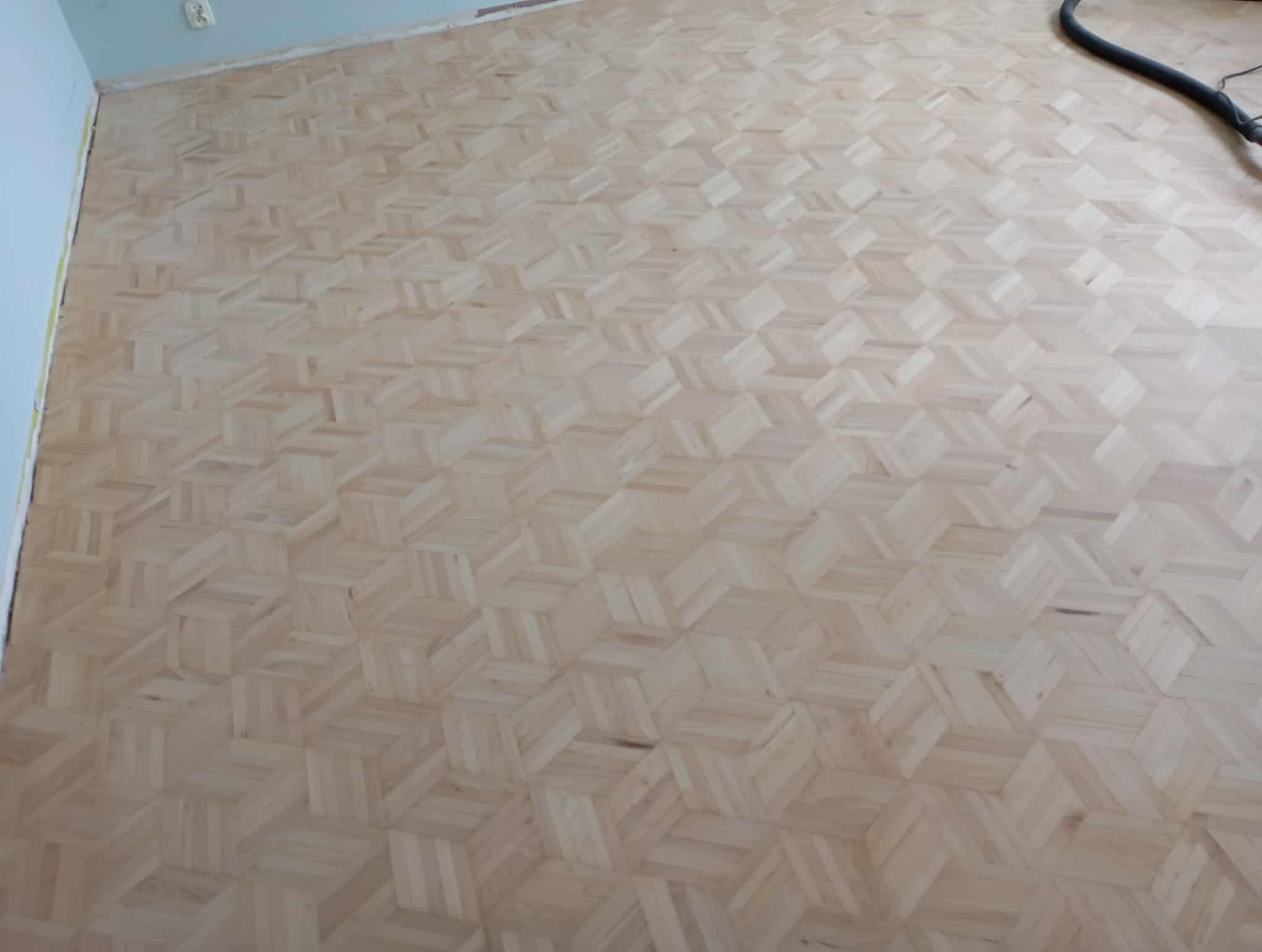 Jasny parkiet jesionowy ułożony w mozaikę 3D, po wstępnym etapie szlifowania. W tle widoczny fragment węża od cykliniarki.