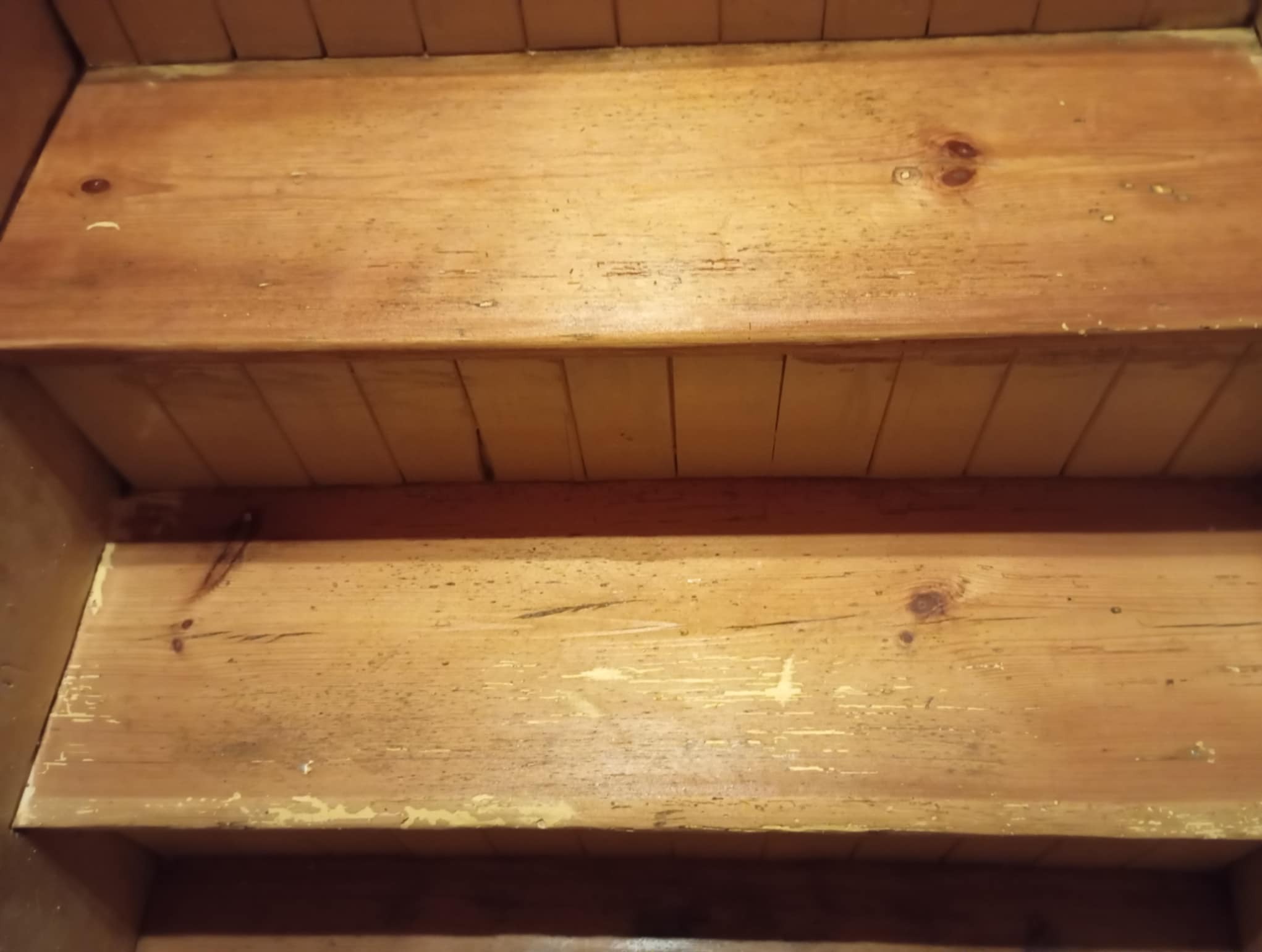 Drewniane schody z widocznymi śladami starej farby, przygotowane do renowacji. Widoczne sęki i naturalna struktura drewna sosnowego.