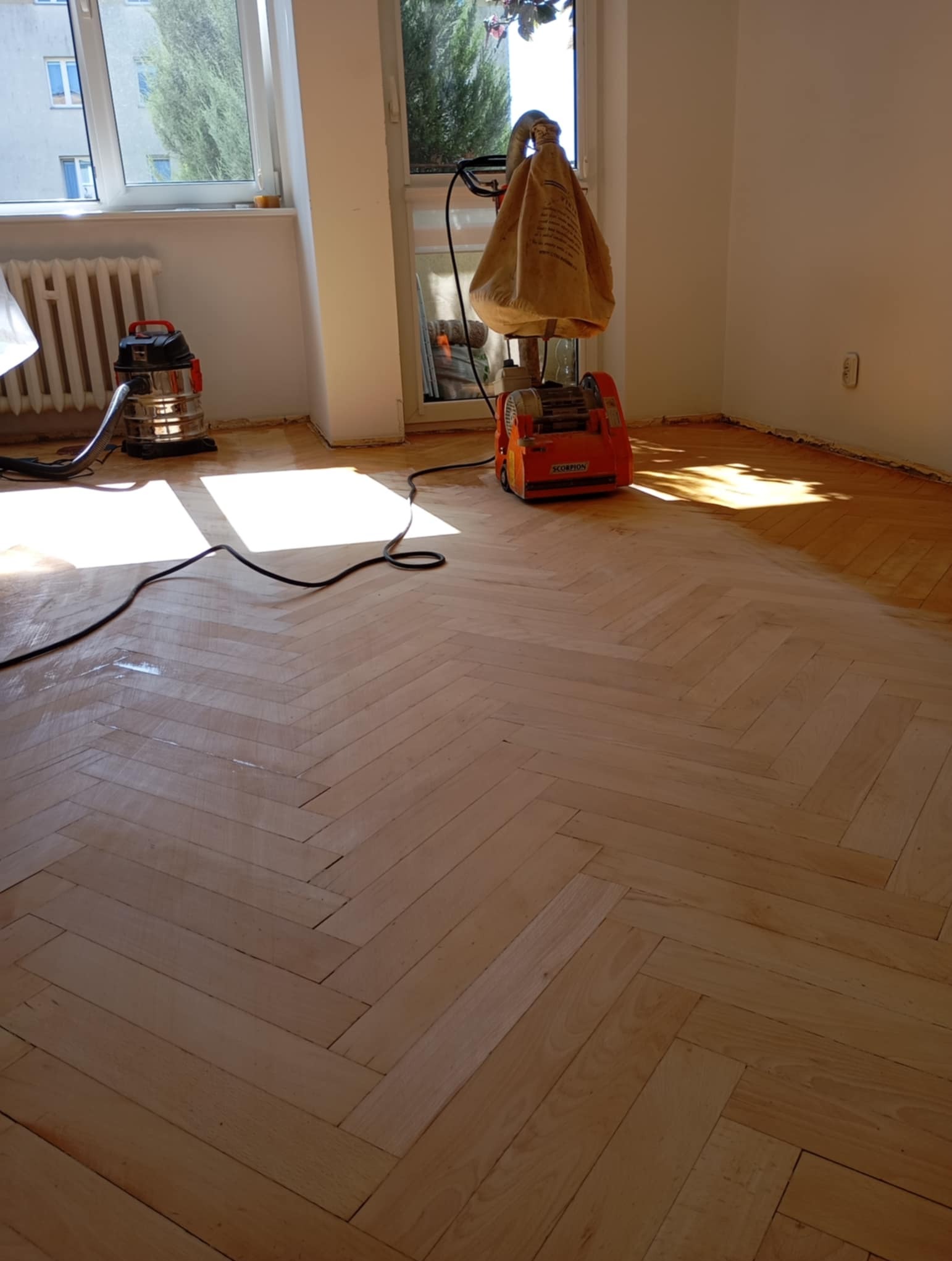 Proces renowacji parkietu dębowego w jodełkę. Widoczna cykliniarka Scorpion i odkurzacz. Część podłogi już odnowiona, jaśniejsza, reszta ciemniejsza.