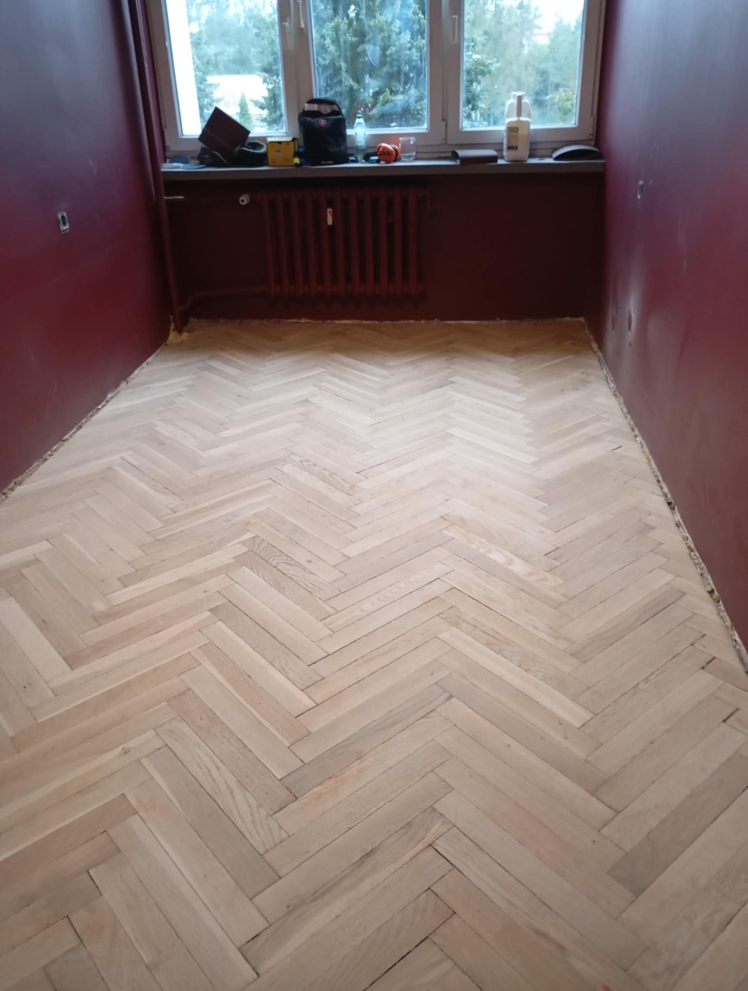 Jasny parkiet dębowy ułożony w jodełkę po cyklinowaniu. Widok na pokój z czerwonymi ścianami i oknem, na parapecie narzędzia i akcesoria.