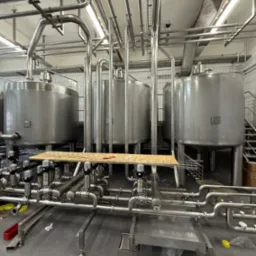Wnętrze przemysłowe z trzema dużymi, metalowymi zbiornikami i rozbudowaną siecią rur. Widoczne elementy instalacji technologicznej w hali produkcyjnej.
