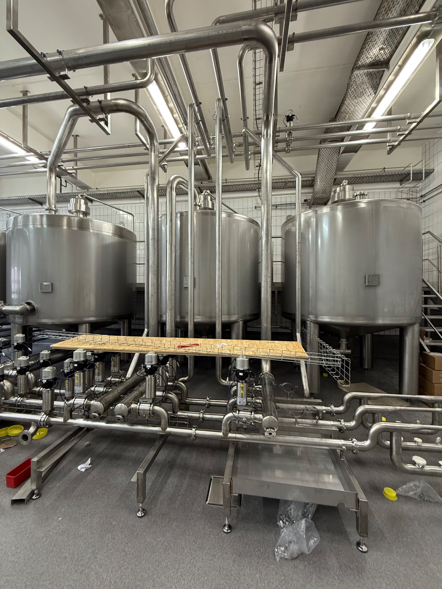 Wnętrze przemysłowe z trzema dużymi, metalowymi zbiornikami i rozbudowaną siecią rur. Widoczne elementy instalacji technologicznej w hali produkcyjnej.