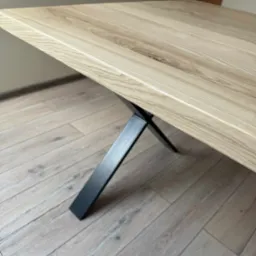 Stół z blatem z jasnego drewna i czarną, metalową nogą w kształcie litery X, ustawiony na podłodze z paneli. Widok z niskiego kąta, ukazujący strukturę drewna.