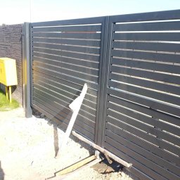 Dominika Zborowska - Nowoczesna, przesuwna brama wjazdowa z poziomymi panelami w kolorze grafitowym. Widoczne ślady montażu i żółta skrzynka gazowa obok ogrodzenia.