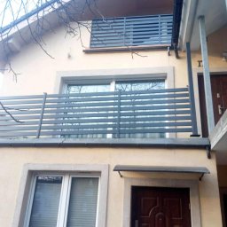 Dominika Zborowska - Nowoczesna, pozioma balustrada balkonowa z metalowych profili, szara, zamontowana na budynku o jasnej elewacji. Widok z dołu, fragment drzewa w kadrze.