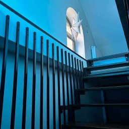 Dominika Zborowska - Nowoczesna balustrada schodowa z pionowych, czarnych prętów, oświetlona niebieskim światłem, z widocznym oknem łukowym i dekoracją w kształcie gwiazdy.