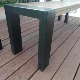 Dominika Zborowska - Metalowa noga stołu na drewnianym tarasie. Noga w kształcie litery U, pomalowana na czarno, mocowana do drewnianego blatu. Widoczne deski tarasowe i zieleń w tle.