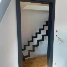 Dominika Zborowska - Nowoczesne, minimalistyczne schody metalowe w czarnej ramie, widoczne z perspektywy wejścia. Drewniana podłoga. Widoczne niedokończone prace elektryczne.