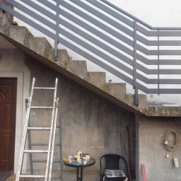 Dominika Zborowska - Szara, pozioma balustrada metalowa zamontowana na betonowych schodach zewnętrznych. W tle widoczna drabina, stolik z naczyniami i krzesła.