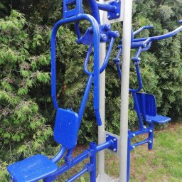 Dominika Zborowska - Kompleksowe, niebieskie urządzenie do ćwiczeń na świeżym powietrzu, z dwoma stanowiskami, na tle zielonych drzew i trawy. Metalowa konstrukcja z siedziskami.