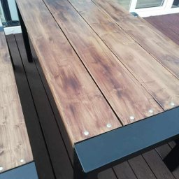 Dominika Zborowska - Drewniany stół ogrodowy z metalowymi nogami, widoczne śruby mocujące deski blatu. Stół na drewnianym tarasie, w tle okno.