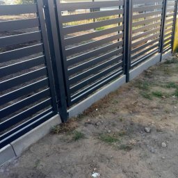 Dominika Zborowska - Nowoczesne ogrodzenie metalowe, poziome panele w kolorze grafitowym, osadzone na betonowej podmurówce. Dom w tle, trawa i ziemia w bliskim planie.