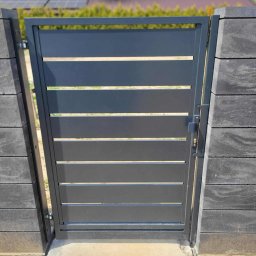 Dominika Zborowska - Nowoczesna, grafitowa furtka z poziomymi panelami, zamontowana między słupkami z szarej kostki. W tle widoczny dom z panelami słonecznymi na dachu.