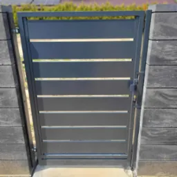 Nowoczesna, grafitowa furtka z poziomymi panelami, zamontowana między słupkami z szarej kostki. W tle widoczny dom z panelami słonecznymi na dachu.