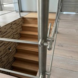 Dominika Zborowska - Nowoczesna balustrada nierdzewna przy drewnianych schodach, z widocznymi detalami mocowań i fakturą podłogi. Minimalistyczny design, jasne wnętrze.
