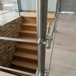 Nowoczesna balustrada nierdzewna przy drewnianych schodach, z widocznymi detalami mocowań i fakturą podłogi. Minimalistyczny design, jasne wnętrze.