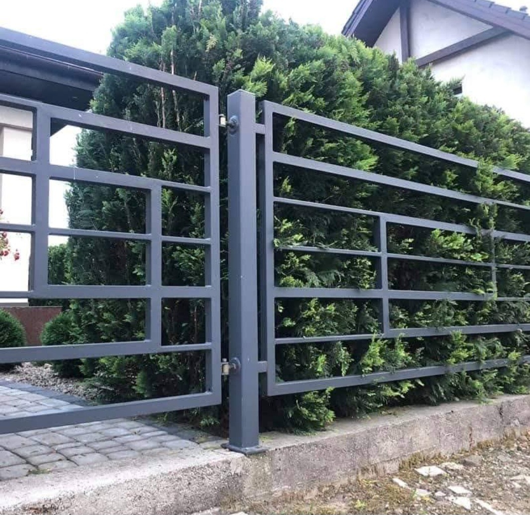 Nowoczesna, grafitowa brama wjazdowa z metalowymi przęsłami na tle zieleni, widoczna kostka brukowa i fragment dachu budynku. Minimalistyczny design.